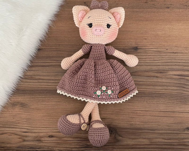 A charming handmade crocheted piggy toy dressed in a brown dress with embroidered flowers, customizable with a name on a leather label. Perfect for baby showers and personalized gifts.