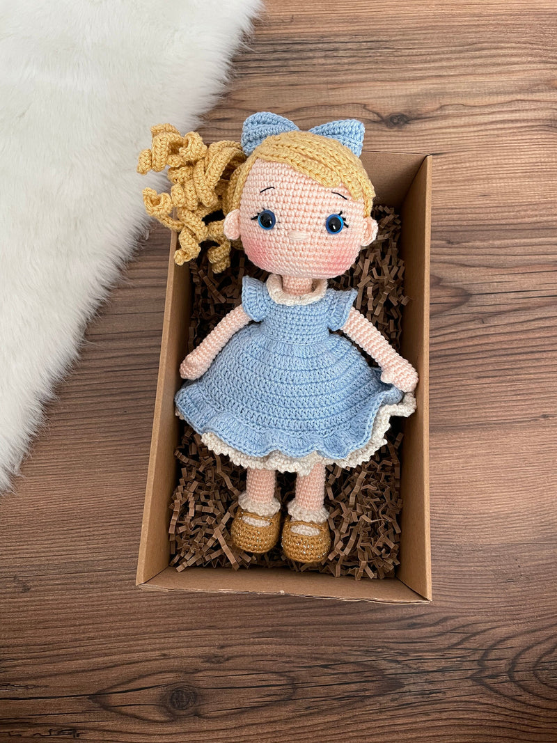 Handmade Blue Nina Doll with blonde hair, wearing a blue dress and a matching bow, made with 100% cotton yarn, customizable with a name on a leather label. Lying inside a gift box.