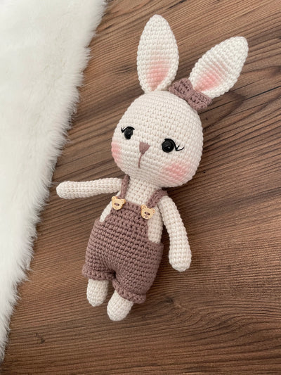 A mink color handmade crocheted Overall Girl Bunny Rabbit Toy with a cute bow and overalls. This amigurumi bunny doll is customizable with a name on a leather label, making it an ideal choice for nursery decor and personalized gifts.