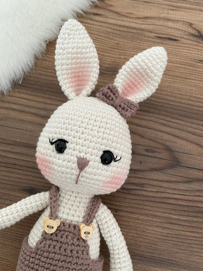 A mink color handmade crocheted Overall Girl Bunny Rabbit Toy with a cute bow and overalls. This amigurumi bunny doll is customizable with a name on a leather label, making it an ideal choice for nursery decor and personalized gifts.