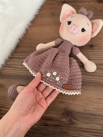 A charming handmade crocheted piggy toy dressed in a brown dress with embroidered flowers, customizable with a name on a leather label. Perfect for baby showers and personalized gifts.