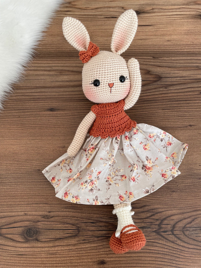 Handmade crocheted bunny rabbit toy dressed in a brick-colored top and floral fabric dress. This amigurumi bunny is customizable with a name on a leather label, perfect for personalized baby gifts.