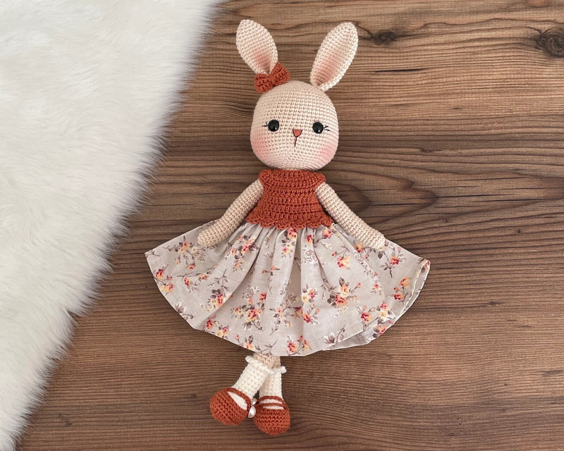 Handmade crocheted bunny rabbit toy dressed in a brick-colored top and floral fabric dress. This amigurumi bunny is customizable with a name on a leather label, perfect for personalized baby gifts.