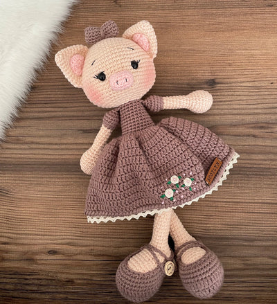 A charming handmade crocheted piggy toy dressed in a brown dress with embroidered flowers, customizable with a name on a leather label. Perfect for baby showers and personalized gifts.