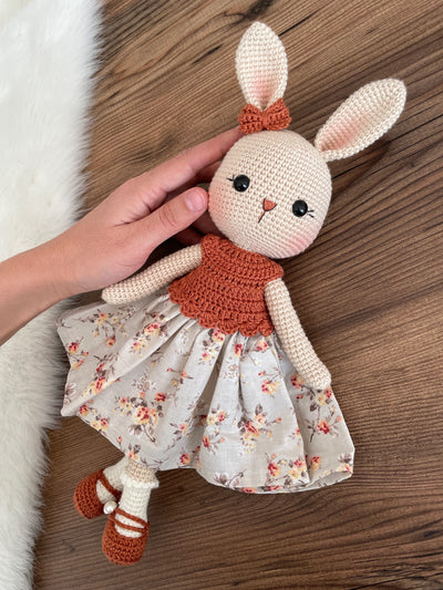 Handmade crocheted bunny rabbit toy dressed in a brick-colored top and floral fabric dress. This amigurumi bunny is customizable with a name on a leather label, perfect for personalized baby gifts.