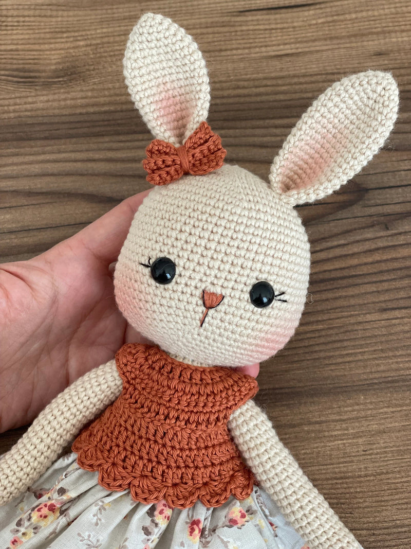 Handmade crocheted bunny rabbit toy dressed in a brick-colored top and floral fabric dress. This amigurumi bunny is customizable with a name on a leather label, perfect for personalized baby gifts.