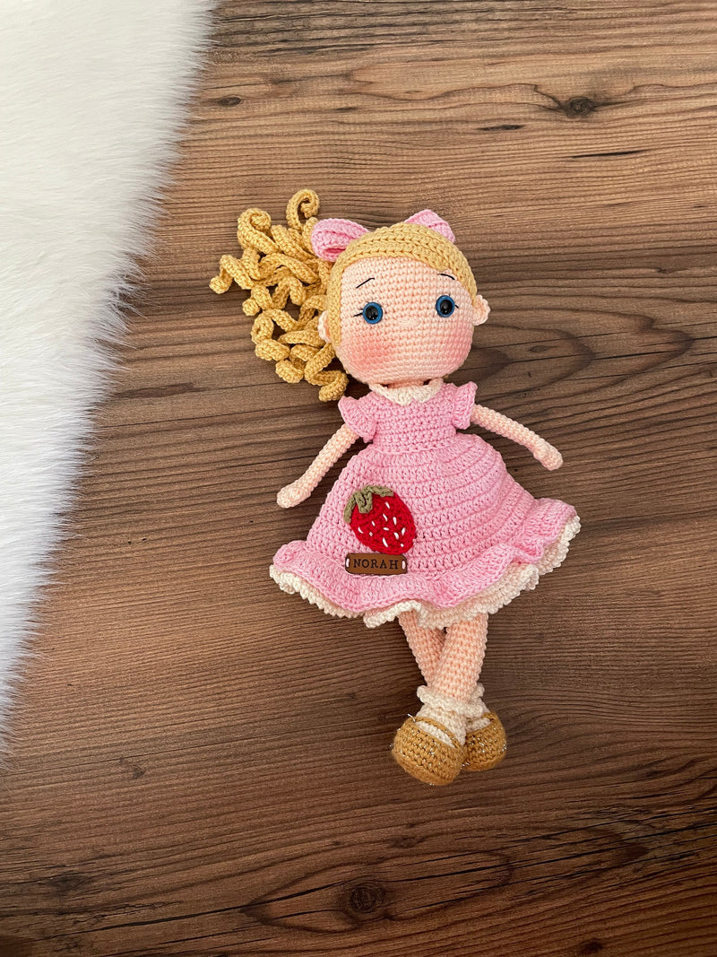 Handmade Pink Nina Doll with blonde hair, wearing a pink dress with a strawberry detail, made with 100% cotton yarn, customizable with a name on a leather label.