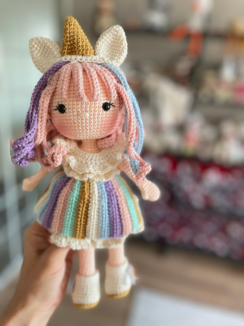 A handmade crocheted Unicorn Girl Doll with pastel rainbow dress, pink curly hair, and a golden horn. This amigurumi toy is customizable with a name on a leather label, perfect for nursery decor and personalized gifts.