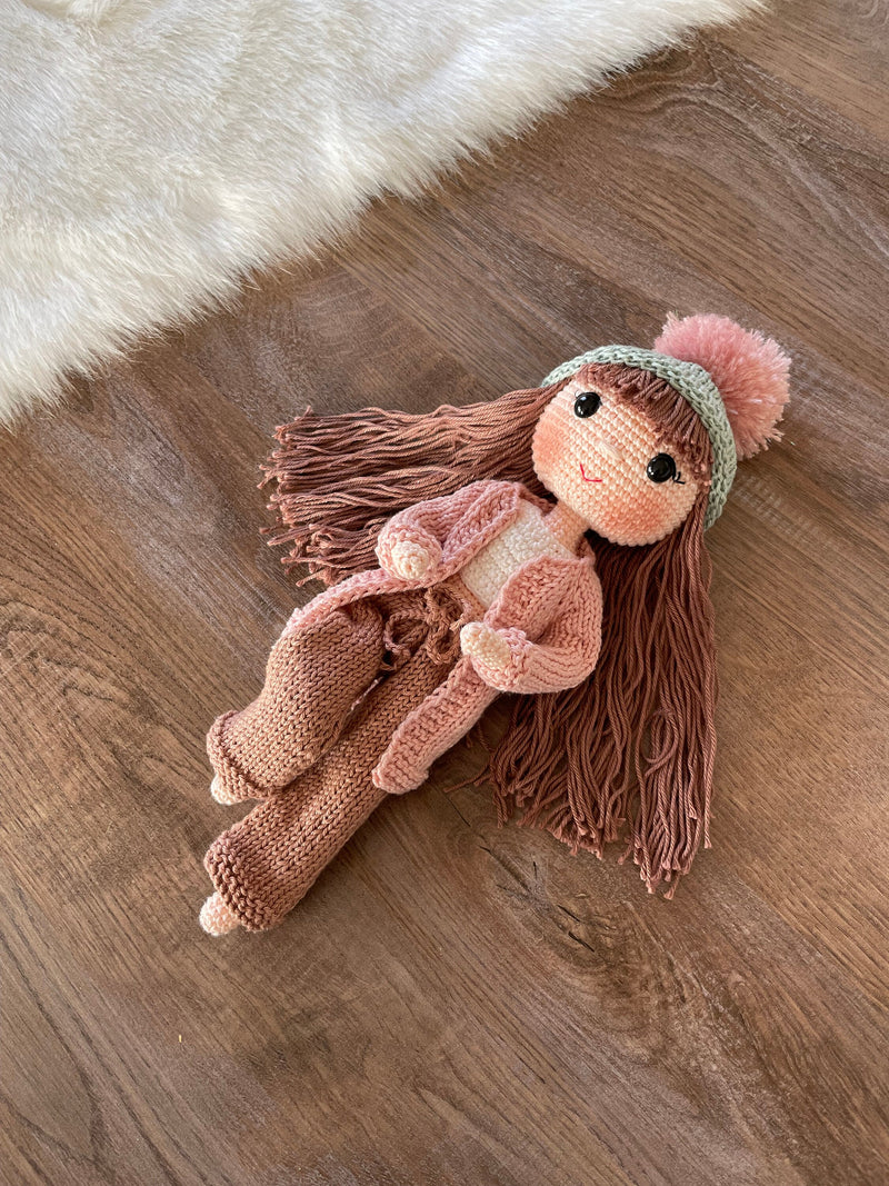 A handcrafted Pink Cardigan Cool Girl Doll with long brown hair, wearing a stylish pink cardigan and beanie, crocheted using the amigurumi technique, and personalized with a custom name on a leather label.