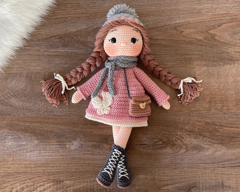 Handmade Pink Winter Girl Doll in a pink coat, gray scarf, and hat with braids, made with 100% cotton yarn, customizable with a name on a leather label.