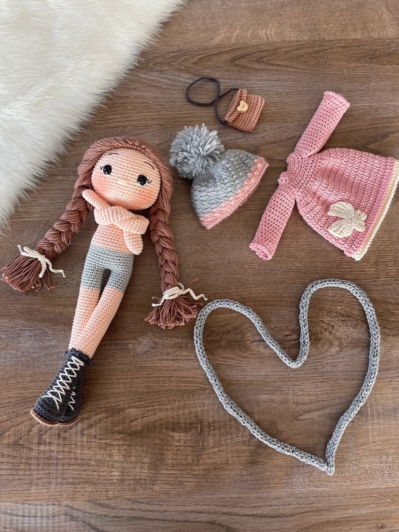 Handmade Pink Winter Girl Doll in a pink coat, gray scarf, and hat with braids, made with 100% cotton yarn, customizable with a name on a leather label.