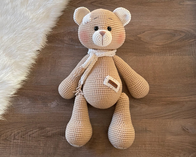 A handmade beige teddy bear toy crafted with the amigurumi technique, featuring a soft texture, a personalized leather name patch, and a cute, cuddly design, perfect for personalized gifts.