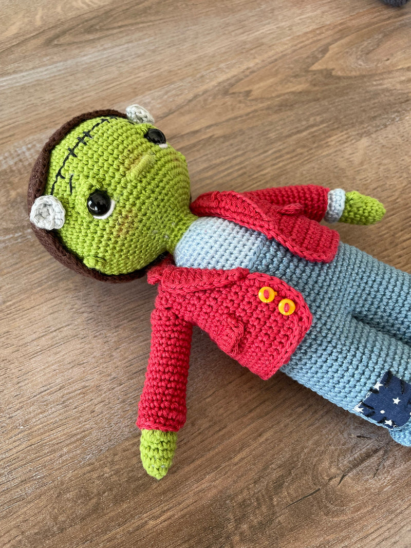 A handmade crochet Frankenstein toy featuring green skin, a red jacket, and patchwork pants. The doll is made using the amigurumi technique and is personalized with a custom name on a leather label.