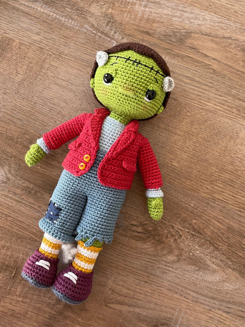 A handmade crochet Frankenstein toy featuring green skin, a red jacket, and patchwork pants. The doll is made using the amigurumi technique and is personalized with a custom name on a leather label.