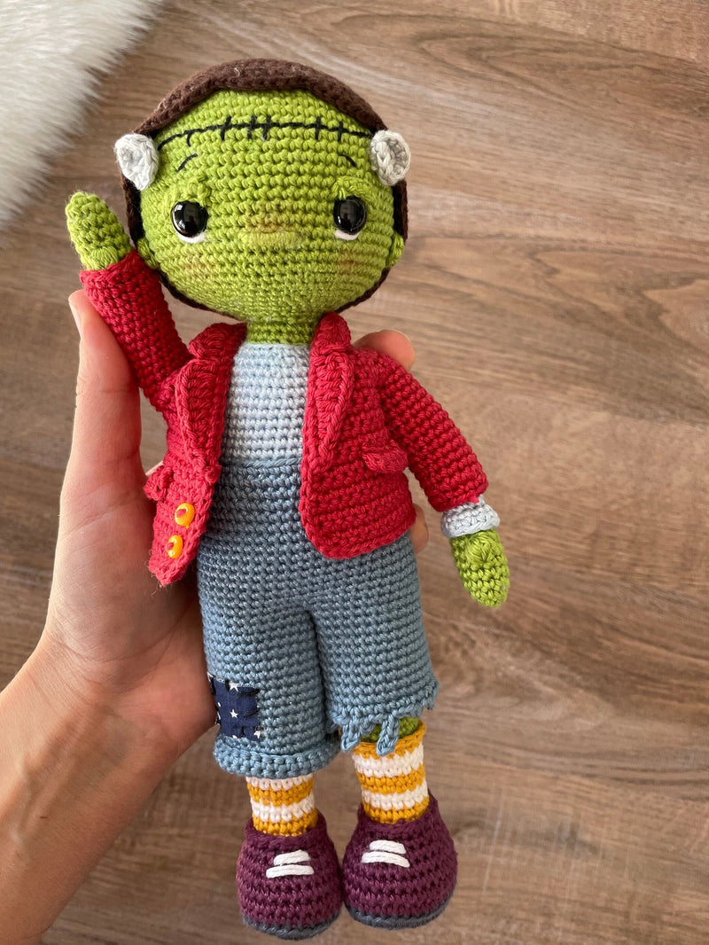 A handmade crochet Frankenstein toy featuring green skin, a red jacket, and patchwork pants. The doll is made using the amigurumi technique and is personalized with a custom name on a leather label.