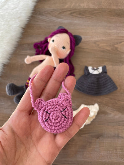 A handcrafted Cat Girl Doll with rich purple hair, wearing a dark dress with cat ears and holding a pink cat-shaped purse. The doll is crocheted using the amigurumi technique and personalized with a custom name on a leather label.