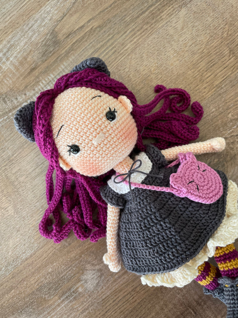 A handcrafted Cat Girl Doll with rich purple hair, wearing a dark dress with cat ears and holding a pink cat-shaped purse. The doll is crocheted using the amigurumi technique and personalized with a custom name on a leather label.