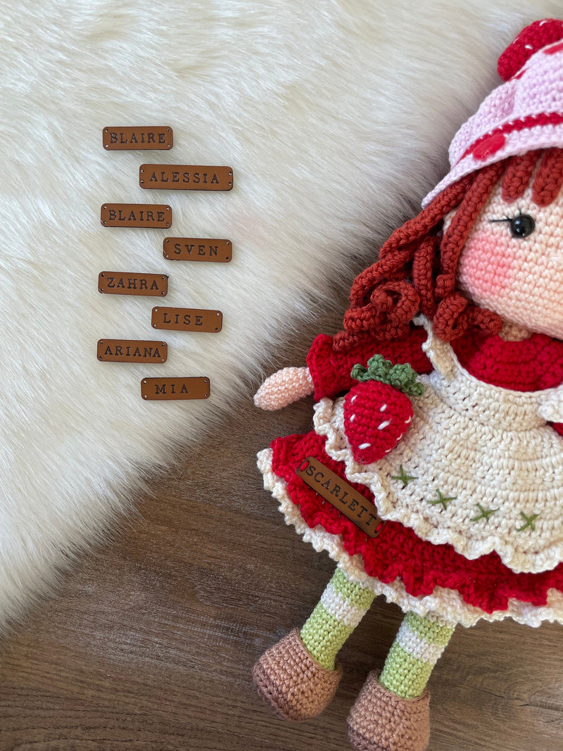 Custom leather name tags with various names lying on next to a strawberry shortcake crochet doll.