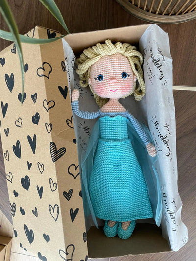 Handmade crocheted amigurumi dolls of Elsa and Anna, inspired by Frozen. Detailed with colorful outfits, perfect for custom name personalization or gifting. Lying inside a gift box.